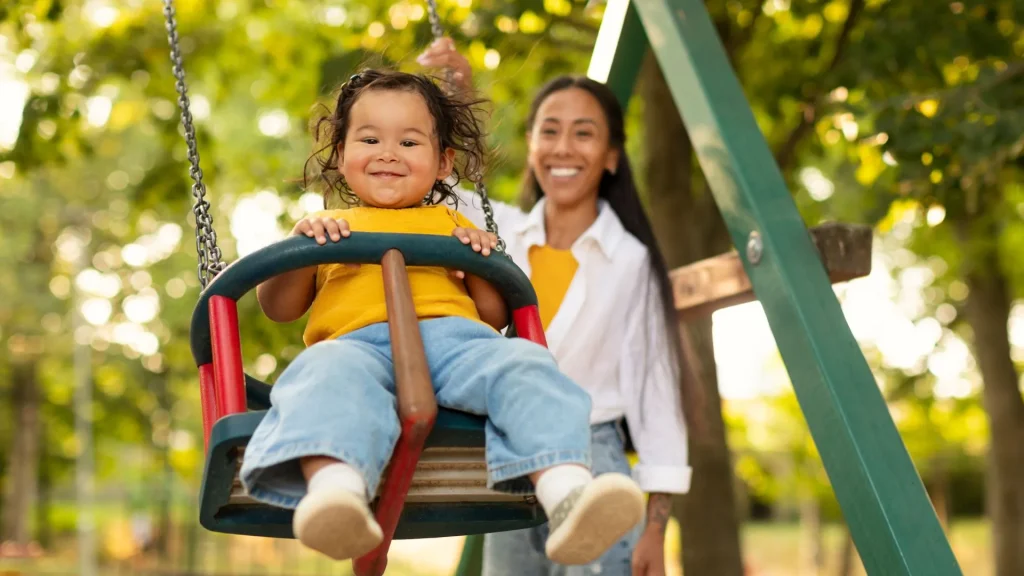 Uma criança pequena sorridente, com cabelo esvoaçado e camiseta amarela, está sentada em um balanço de parquinho (swing set) em primeiro plano. Uma mulher, presumivelmente a mãe, de cabelo escuro preso e camisa branca, está em segundo plano, empurrando o balanço. Ambas parecem alegres, e o cenário é um parque verde e ensolarado com árvores.
