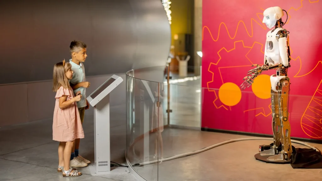 Duas crianças (um menino e uma menina) estão em um museu de ciências, observando um robô humanoide de metal em exibição. O menino está de camisa polo e a menina está de vestido rosa-claro. Elas estão em frente a um painel de vidro curvo, olhando para o robô. O fundo é uma parede vermelha com um desenho de engrenagem em amarelo e laranja.