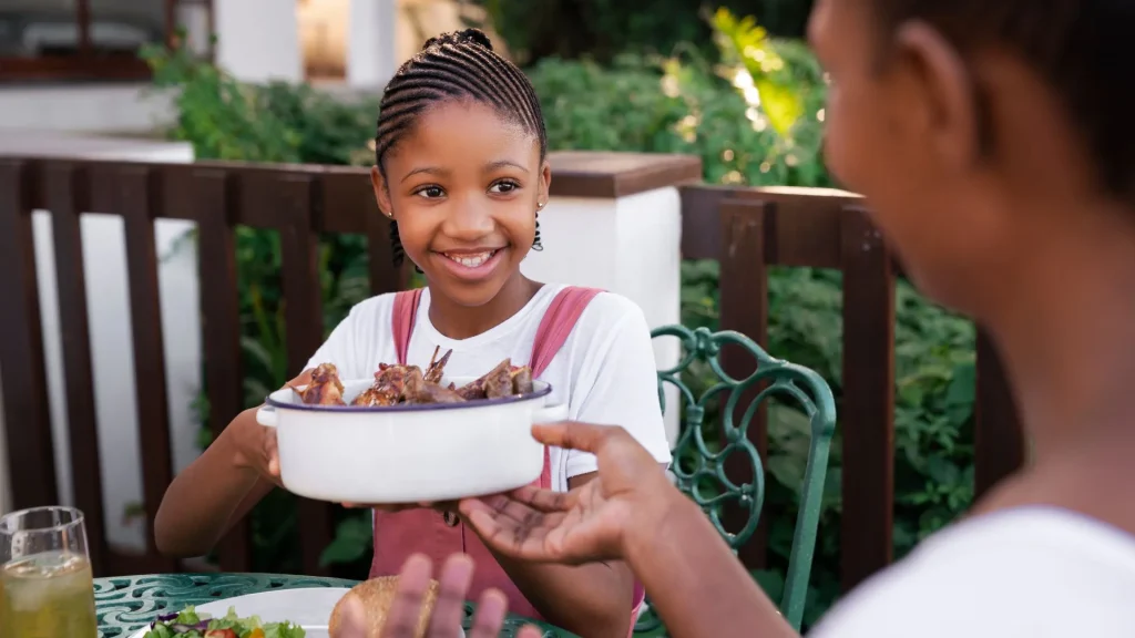 Menina sorridente de tranças, vestindo um macacão rosa e camiseta branca, está entregando uma travessa branca cheia de carne assada ou grelhada para uma pessoa adulta (fora de foco) durante uma refeição ao ar livre. Há uma mesa com um prato de salada e um copo de bebida.