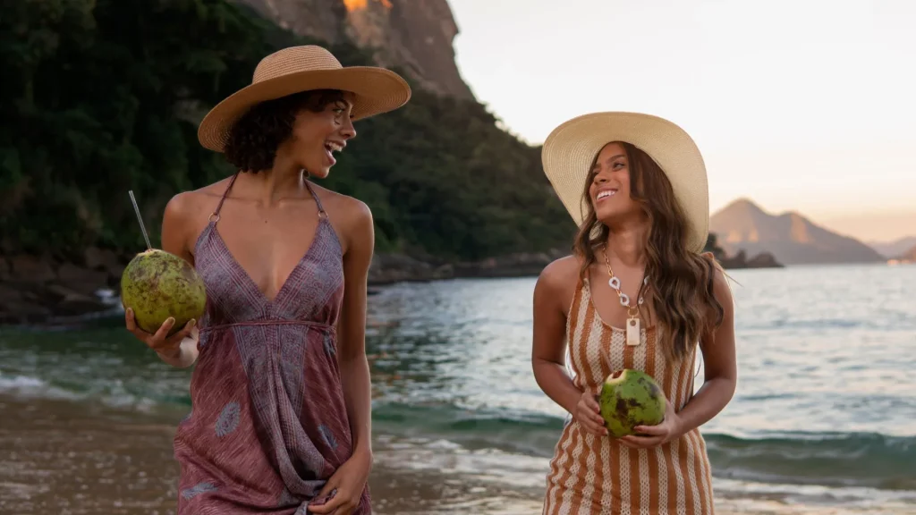 Duas mulheres sorridentes, de chapéus de aba larga e vestidos de verão, caminham na areia da praia ao pôr do sol, segurando cocos verdes. Ao fundo, uma paisagem montanhosa e o mar sereno.