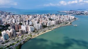 Vista aérea e panorâmica do horizonte de Florianópolis, Santa Catarina, Brasil. A imagem mostra uma densa área urbana de prédios de diversos tamanhos e cores claras, que se estende ao longo de uma ampla avenida costeira. A avenida é ladeada por uma faixa de areia estreita e pelo mar, com águas em tons de azul-turquesa e verde-azulado. Ao fundo, pode-se ver o restante da cidade, colinas cobertas de vegetação, e uma ponte que liga a ilha ao continente. O céu é azul com algumas nuvens brancas.