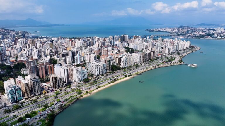 Vista aérea e panorâmica do horizonte de Florianópolis, Santa Catarina, Brasil. A imagem mostra uma densa área urbana de prédios de diversos tamanhos e cores claras, que se estende ao longo de uma ampla avenida costeira. A avenida é ladeada por uma faixa de areia estreita e pelo mar, com águas em tons de azul-turquesa e verde-azulado. Ao fundo, pode-se ver o restante da cidade, colinas cobertas de vegetação, e uma ponte que liga a ilha ao continente. O céu é azul com algumas nuvens brancas.