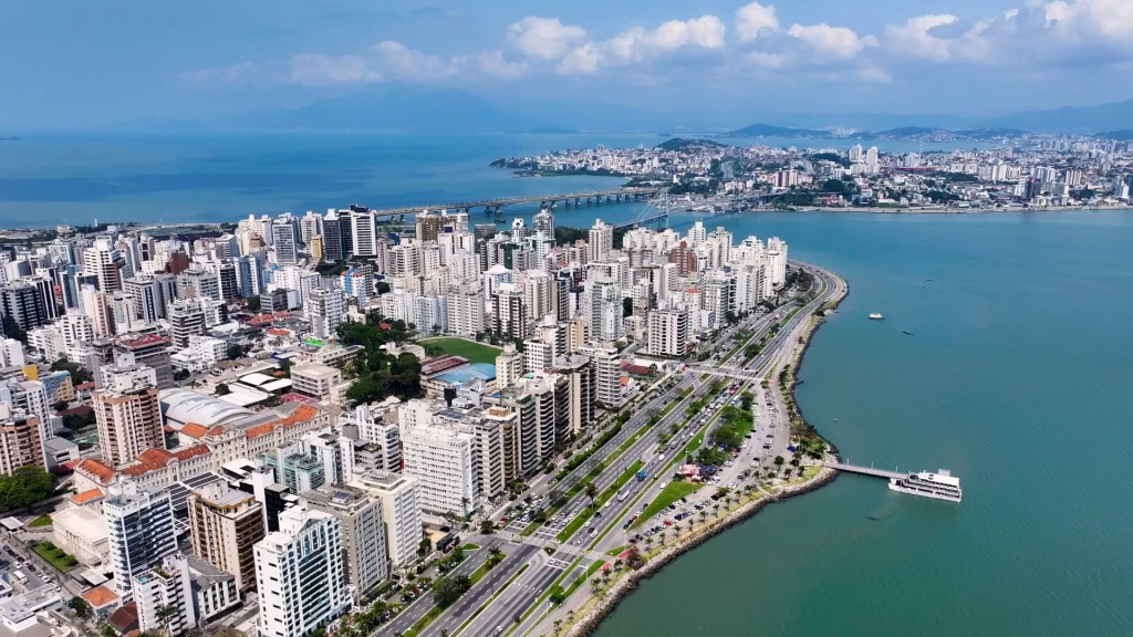 Vista aérea do centro de Florianópolis, Santa Catarina, mostrando a densa área urbana e a orla marítima. Prédios altos e modernos se estendem ao longo da costa, separados do mar por uma avenida expressa. A paisagem costeira forma uma curva que segue o contorno da ilha. Ao fundo, a Ponte Hercílio Luz é visível, ligando a ilha ao continente, e montanhas cobertas por neblina ou nuvens se erguem no horizonte. O mar e o céu estão em tons de azul com nuvens brancas.