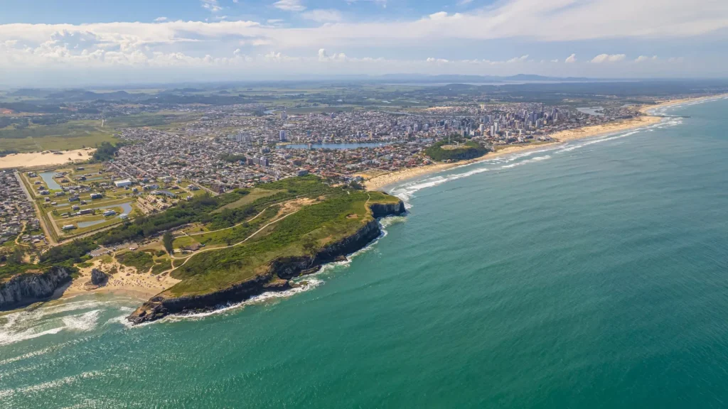 Vista aérea da cidade de Torres, Rio Grande do Sul, mostrando a junção da paisagem urbana com a natureza única do litoral gaúcho. Em primeiro plano, o mar de cor verde-azulada domina a cena. À esquerda, uma das famosas formações rochosas (morros ou falésias) de Torres avança no mar, coberta por vegetação baixa e cercada por pequenas praias de areia. A linha costeira se estende para a direita, revelando uma longa faixa de praia e a área urbana densa da cidade. O horizonte ao fundo é plano, característico do litoral do estado.