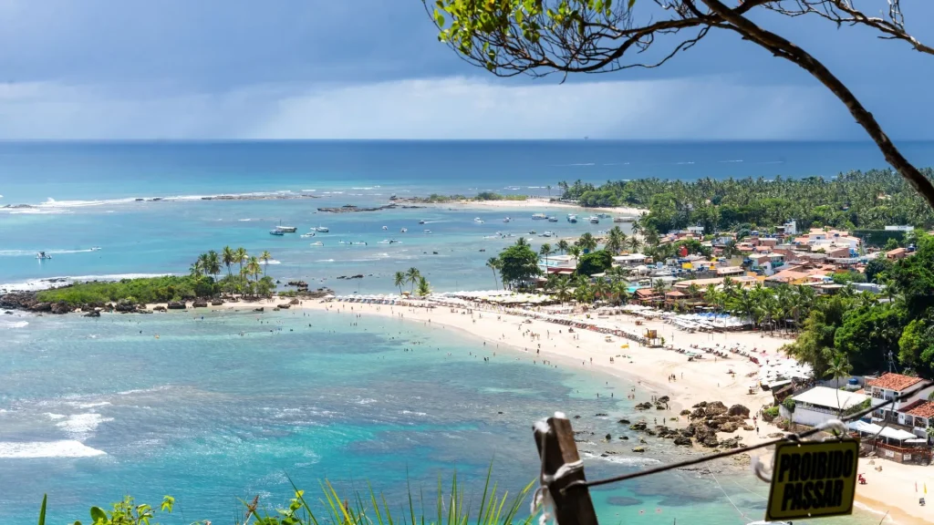 Vista panorâmica de Morro de São Paulo, Bahia, com foco na orla e nas praias. A imagem mostra a Segunda Praia, com areia clara, cheia de pessoas e barracas coloridas. O mar, de um azul-turquesa vibrante, possui águas calmas e rasas, repletas de barcos ancorados. Na esquerda, há um pequeno banco de areia com coqueiros. A área urbana da vila se estende por trás da praia, envolta por vegetação. Em primeiro plano, no canto inferior direito, há uma placa de madeira em português indicando "PROIBIDO PASSAR".