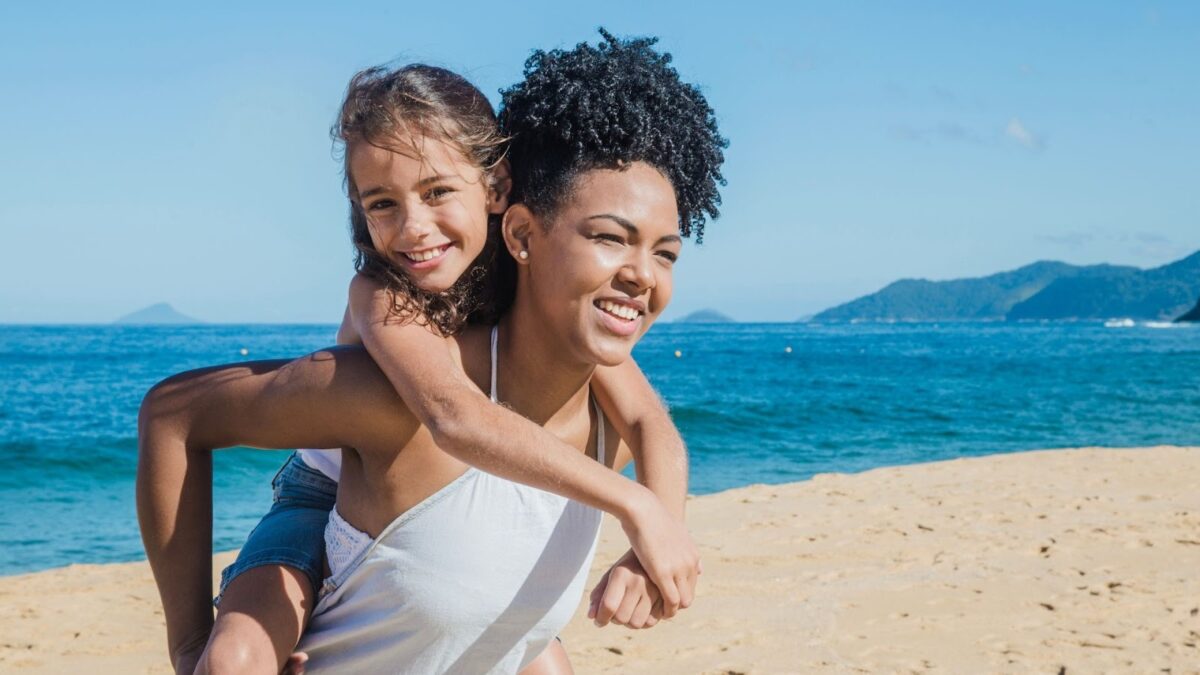 Mãe e filha sorrindo e se divertindo na praia. A mãe, de cabelo cacheado, está carregando a filha nas costas. A filha, que sorri e olha para frente, está com os braços ao redor do pescoço da mãe. O fundo é uma paisagem de praia com areia dourada, mar azul e morros ao longe.