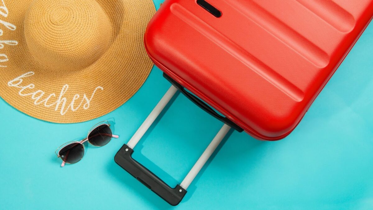 Mala de viagem rígida vermelha com a alça estendida, óculos de sol de armação rosa e dourada e um chapéu de palha com a palavra 'beaches' (praias) bordada em branco, tudo disposto sobre um fundo azul-turquesa. O cenário sugere férias de verão e viagem.