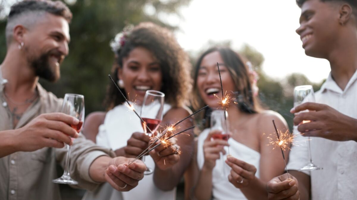 Um grupo de quatro jovens adultos, dois homens e duas mulheres, sorrindo e celebrando ao ar livre. Eles seguram taças com uma bebida avermelhada (possivelmente vinho ou espumante) e pequenos fogos de artifício (sparklers) acesos, que estão soltando faíscas. As mulheres estão vestindo roupas brancas, e os homens usam camisas claras. A cena transmite alegria e celebração, sugerindo uma festa ou um evento especial, como o Ano Novo.