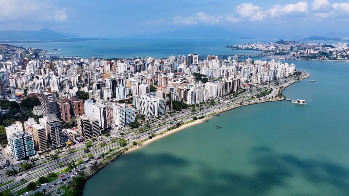 Vista aérea e panorâmica do horizonte de Florianópolis, Santa Catarina, Brasil. A imagem mostra uma densa área urbana de prédios de diversos tamanhos e cores claras, que se estende ao longo de uma ampla avenida costeira. A avenida é ladeada por uma faixa de areia estreita e pelo mar, com águas em tons de azul-turquesa e verde-azulado. Ao fundo, pode-se ver o restante da cidade, colinas cobertas de vegetação, e uma ponte que liga a ilha ao continente. O céu é azul com algumas nuvens brancas.
