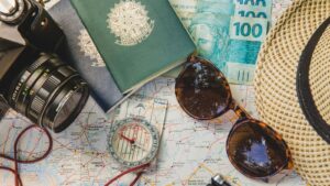 Uma foto aérea exibe itens essenciais para viagem dispostos sobre um mapa aberto da América do Sul. No centro, há dois passaportes brasileiros de capa verde, parcialmente cobertos por notas de Real (moeda brasileira) no valor de R$100. À direita, um chapéu de palha e um par de óculos de sol escuros. Um compasso e um transferidor estão sobre o mapa, e um pedaço de barbante vermelho de bússola guia a visão. No canto esquerdo, uma câmera fotográfica analógica escura compõe a cena, sugerindo planejamento de viagem e aventura.