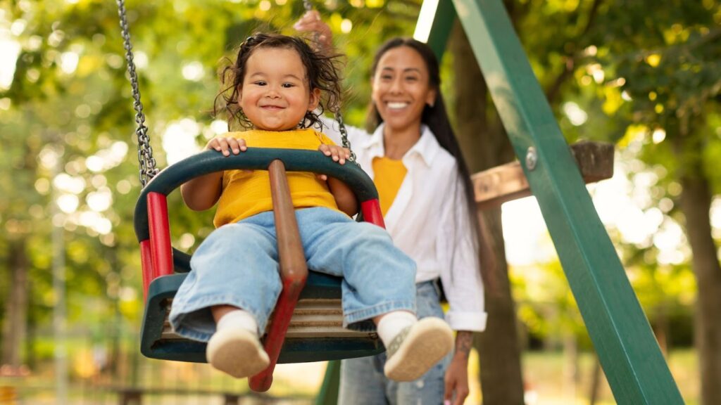 Uma criança pequena sorridente, com cabelo esvoaçado e camiseta amarela, está sentada em um balanço de parquinho (swing set) em primeiro plano. Uma mulher, presumivelmente a mãe, de cabelo escuro preso e camisa branca, está em segundo plano, empurrando o balanço. Ambas parecem alegres, e o cenário é um parque verde e ensolarado com árvores.