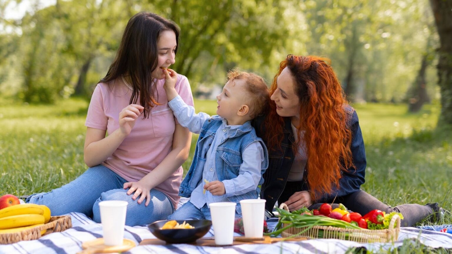 Duas mulheres e uma criança pequena estão fazendo um piquenique em um parque em um dia ensolarado. A mulher da esquerda, de cabelo escuro e camiseta rosa, está sorrindo enquanto a criança, no centro, de colete jeans azul, estende a mão para lhe dar um pedaço de comida. A mulher da direita, de cabelo ruivo vibrante e jaqueta jeans, olha para a criança com um sorriso. Elas estão sentadas em um cobertor xadrez com frutas, vegetais e copos de papel na frente delas, cercadas por grama verde e árvores desfocadas.