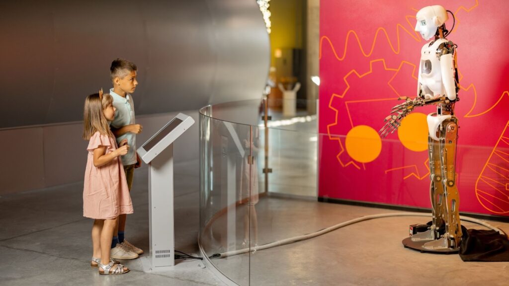 Duas crianças (um menino e uma menina) estão em um museu de ciências, observando um robô humanoide de metal em exibição. O menino está de camisa polo e a menina está de vestido rosa-claro. Elas estão em frente a um painel de vidro curvo, olhando para o robô. O fundo é uma parede vermelha com um desenho de engrenagem em amarelo e laranja.