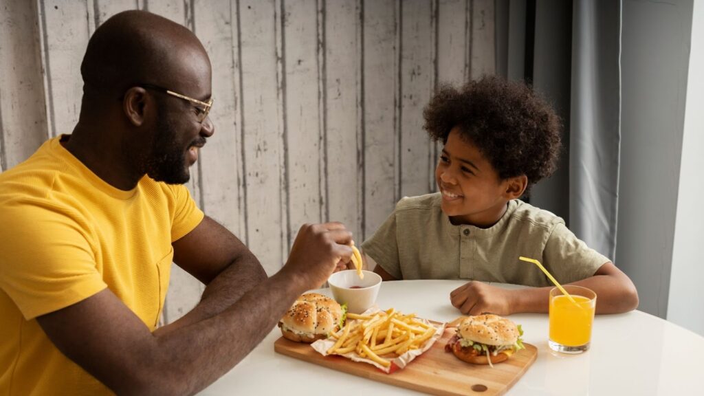 Um homem adulto sorridente, de óculos e camisa polo amarela, está sentado à esquerda de uma mesa branca. Ele está mergulhando uma batata frita em um pequeno pote de ketchup que está no centro da mesa. À direita, um menino com cabelo crespo e camisa verde-oliva sorri, olhando para o homem. Entre eles, em uma tábua de madeira, há duas porções de hambúrguer e uma grande porção de batatas fritas. Há também um copo de suco de laranja com um canudo amarelo.
