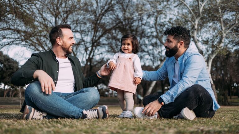 Uma família composta por dois pais e uma filha pequena sentada em um gramado de um parque. Os dois homens estão sentados de pernas cruzadas no chão, sorrindo e olhando para a criança que está no centro, de pé, usando um vestido rosa. O homem à esquerda veste uma camisa branca sob uma jaqueta verde escura. O homem à direita veste uma camisa social azul clara e tem barba. O fundo é de árvores desfocadas.