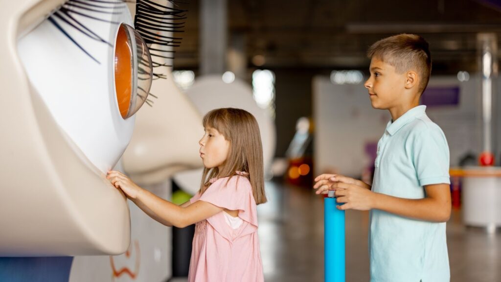 Duas crianças estão em um museu ou exposição de ciências, observando uma grande maquete de um olho humano em primeiro plano. A menina, à esquerda, com cabelos castanhos e usando um vestido rosa pálido, toca a lateral do modelo do olho, que tem uma íris laranja brilhante e cílios grandes. O menino, à direita, usa uma camisa polo azul clara e observa o modelo de perfil.