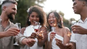 Um grupo de quatro jovens adultos, dois homens e duas mulheres, sorrindo e celebrando ao ar livre. Eles seguram taças com uma bebida avermelhada (possivelmente vinho ou espumante) e pequenos fogos de artifício (sparklers) acesos, que estão soltando faíscas. As mulheres estão vestindo roupas brancas, e os homens usam camisas claras. A cena transmite alegria e celebração, sugerindo uma festa ou um evento especial, como o Ano Novo.