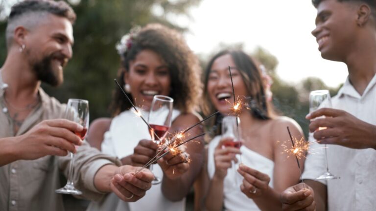 Um grupo de quatro jovens adultos, dois homens e duas mulheres, sorrindo e celebrando ao ar livre. Eles seguram taças com uma bebida avermelhada (possivelmente vinho ou espumante) e pequenos fogos de artifício (sparklers) acesos, que estão soltando faíscas. As mulheres estão vestindo roupas brancas, e os homens usam camisas claras. A cena transmite alegria e celebração, sugerindo uma festa ou um evento especial, como o Ano Novo.