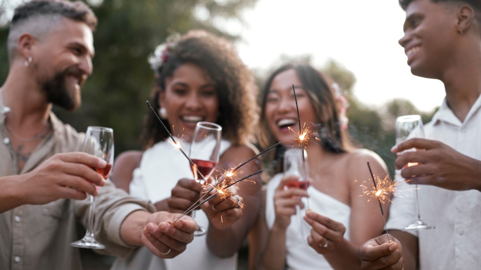 Um grupo de quatro jovens adultos, dois homens e duas mulheres, sorrindo e celebrando ao ar livre. Eles seguram taças com uma bebida avermelhada (possivelmente vinho ou espumante) e pequenos fogos de artifício (sparklers) acesos, que estão soltando faíscas. As mulheres estão vestindo roupas brancas, e os homens usam camisas claras. A cena transmite alegria e celebração, sugerindo uma festa ou um evento especial, como o Ano Novo.