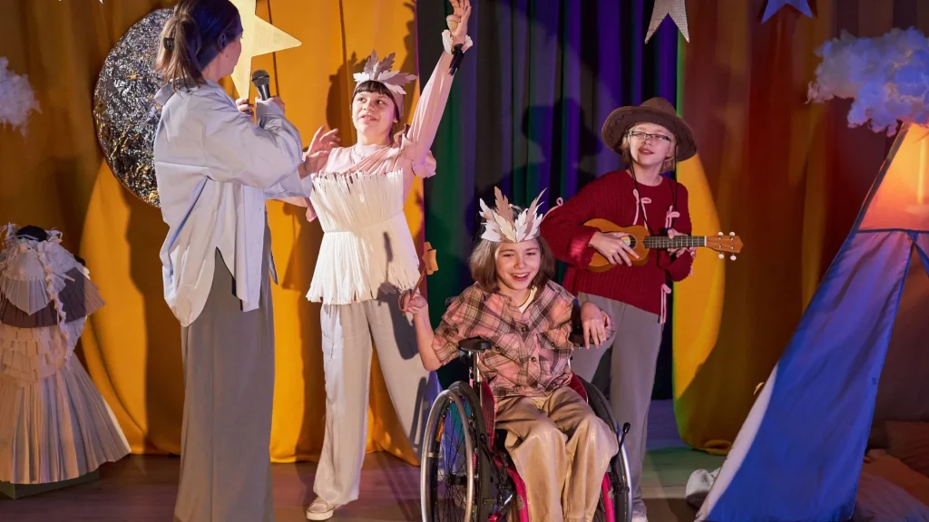 Uma imagem vibrante de um palco com quatro pessoas participando de uma apresentação teatral. A decoração sugere um tema celestial ou de acampamento, com estrelas douradas penduradas, nuvens de algodão e um tecido laranja-avermelhado ao fundo que lembra uma tenda. Da esquerda para a direita: uma mulher adulta, vestida casualmente, segura um microfone e interage com os participantes. No centro, duas meninas estão de pé e sentadas. A menina de pé usa uma roupa branca ou rosa-claro com franjas e uma tiara que parece uma coroa de penas, com os braços erguidos em pose dramática. A menina sentada em uma cadeira de rodas está no primeiro plano, sorridente, vestindo uma camisa xadrez e calças claras, também usando uma coroa de penas. À direita, um menino de chapéu de cowboy, óculos e suéter vermelho toca um ukulele.