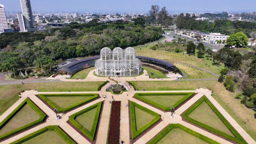 Vista aérea do Jardim Botânico de Curitiba, Paraná. No centro, destaca-se a icônica estufa de metal e vidro em estilo art nouveau com três cúpulas. À frente da estufa, estende-se um jardim em estilo francês com gramados simétricos em formatos geométricos, cercados por sebes verdes e caminhos pavimentados. Ao fundo, uma densa área de mata nativa e a silhueta urbana da cidade sob um céu azul claro.