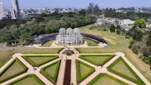 Vista aérea do Jardim Botânico de Curitiba, Paraná. No centro, destaca-se a icônica estufa de metal e vidro em estilo art nouveau com três cúpulas. À frente da estufa, estende-se um jardim em estilo francês com gramados simétricos em formatos geométricos, cercados por sebes verdes e caminhos pavimentados. Ao fundo, uma densa área de mata nativa e a silhueta urbana da cidade sob um céu azul claro.