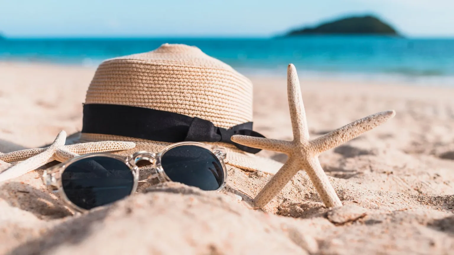 Fotografia em close-up de acessórios de praia sobre a areia clara. No centro, um chapéu de palha com laço preto e um par de óculos de sol com lentes escuras. Duas estrelas-do-mar de cor bege estão posicionadas ao lado do chapéu. O fundo apresenta um mar calmo em tons de turquesa, uma ilha montanhosa e um horizonte ensolarado com efeito levemente desfocado.