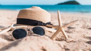 Fotografia em close-up de acessórios de praia sobre a areia clara. No centro, um chapéu de palha com laço preto e um par de óculos de sol com lentes escuras. Duas estrelas-do-mar de cor bege estão posicionadas ao lado do chapéu. O fundo apresenta um mar calmo em tons de turquesa, uma ilha montanhosa e um horizonte ensolarado com efeito levemente desfocado.