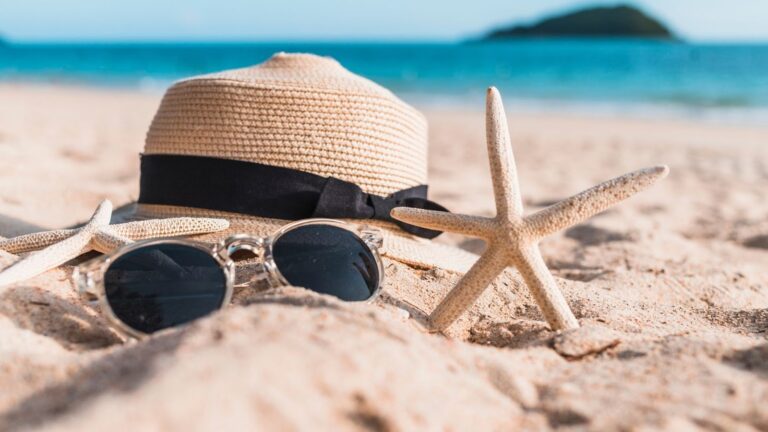 Fotografia em close-up de acessórios de praia sobre a areia clara. No centro, um chapéu de palha com laço preto e um par de óculos de sol com lentes escuras. Duas estrelas-do-mar de cor bege estão posicionadas ao lado do chapéu. O fundo apresenta um mar calmo em tons de turquesa, uma ilha montanhosa e um horizonte ensolarado com efeito levemente desfocado.