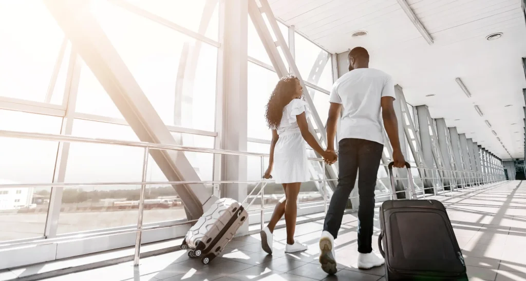 Um casal negro de costas, de mãos dadas, caminha por um corredor amplo e iluminado de um aeroporto. A mulher, à esquerda, usa um vestido branco curto e puxa uma mala de rodinhas bege. O homem, à direita, veste camiseta branca e calça jeans escura, carregando uma mala preta. Eles olham um para o outro enquanto caminham em direção a uma grande janela de vidro que revela uma estrutura metálica externa sob a luz do sol.