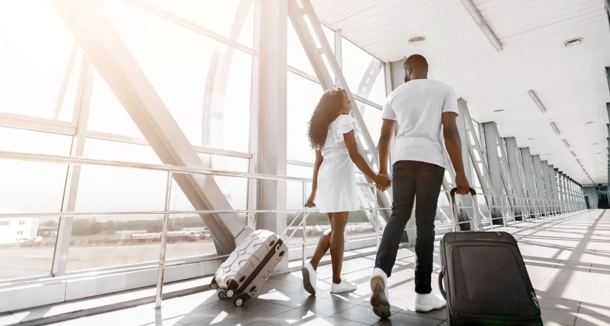 Um casal negro de costas, de mãos dadas, caminha por um corredor amplo e iluminado de um aeroporto. A mulher, à esquerda, usa um vestido branco curto e puxa uma mala de rodinhas bege. O homem, à direita, veste camiseta branca e calça jeans escura, carregando uma mala preta. Eles olham um para o outro enquanto caminham em direção a uma grande janela de vidro que revela uma estrutura metálica externa sob a luz do sol.